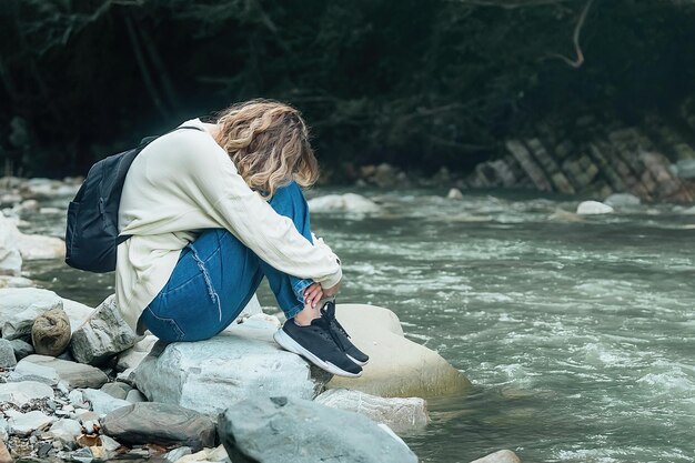 une femme touristique frustrée et fatiguée est assise seule sur la rive d'une rivière de montagne lors d'une promenade dans la nature