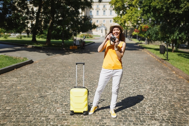 Femme touristique voyageur en chapeau jaune de vêtements décontractés avec valise prenant des photos sur un appareil photo vintage rétro en plein air. Fille voyageant à l'étranger pour voyager en week-end. Mode de vie de voyage touristique.