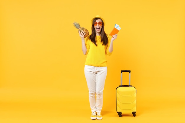 Femme touristique voyageuse excitée au chapeau tenant des billets de passeport, fruit d'ananas frais isolé sur fond jaune orange. Passager voyageant à l'étranger en week-end. Concept de voyage de vol aérien.