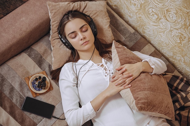 Femme tranquille écoutant de la musique dans des écouteurs et se reposant sur le lit dans la chambre domestique