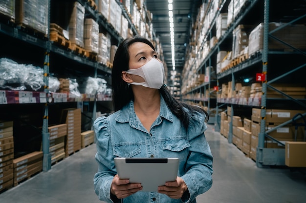 Femme de travail asiatique portant un masque à l'aide d'une tablette numérique contrôle du stock de produits dans l'entrepôt