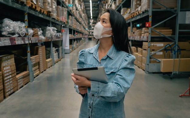 Femme De Travail Asiatique Portant Un Masque à L'aide D'une Tablette Numérique Contrôle Du Stock De Produits Dans L'entrepôt