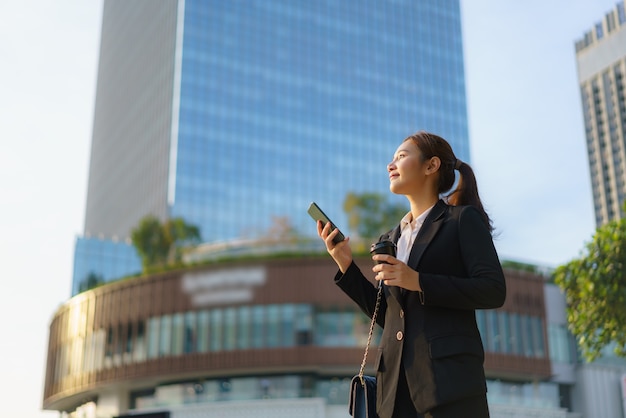 Femme de travail exécutive asiatique tenant une tasse de café et à l'aide d'un téléphone mobile dans la rue avec des immeubles de bureaux en arrière-plan à Bangkok, Thaïlande.