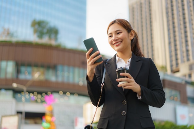 Femme de travail exécutive asiatique tenant une tasse de café et à l'aide d'un téléphone mobile dans la rue avec des immeubles de bureaux en arrière-plan à Bangkok, Thaïlande.