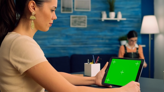 Femme travaillant au bureau à l'aide d'une tablette à écran vert en mode paysage, regardant du contenu en ligne