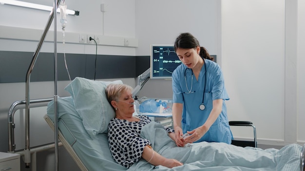Femme travaillant comme infirmière utilisant un oxymètre pour mesurer la saturation en oxygène afin de soigner un patient âgé au lit. Assistant médical mettant l'outil sous la main d'une femme avec un tube à oxygène et un sac d'égouttement IV