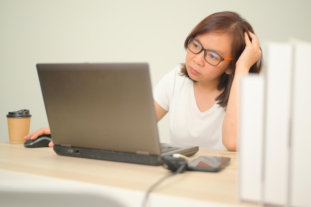 Une femme travaillant à domicile avec un ordinateur portable, portant un t-shirt blanc et des lunettes, les mains sur la souris et la tête, des livres et une tasse à café sur la table, travaille à la maison, bureau à domicile, travail à distance, New Normal.