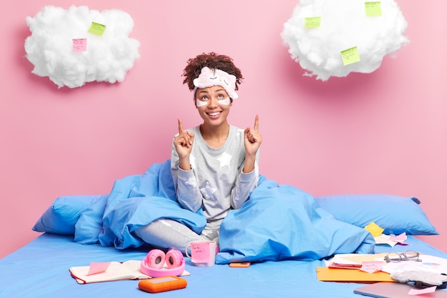 une femme travaille à domicile aime les postes de travail à distance au-dessus sur les nuages porte un masque de sommeil et un pyjama est assise les jambes croisées au lit entourée de notes en papier prépare le projet