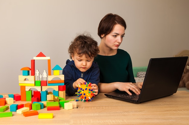 Une femme travaille à la maison avec un enfant. La mère est assise derrière un ordinateur portable pendant que l'enfant joue et fabrique un faux coronavirus