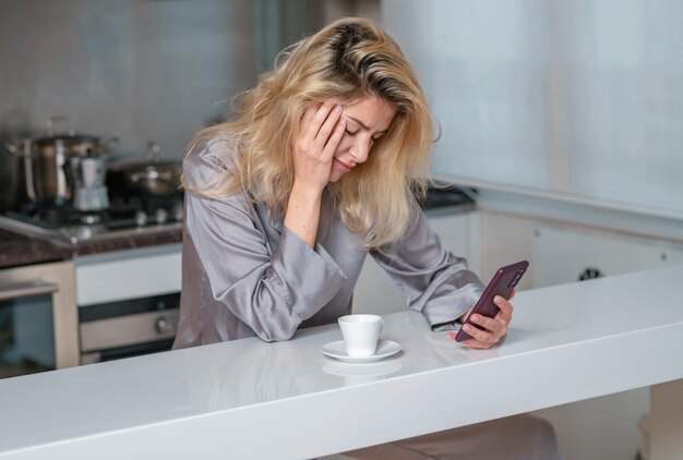 Une femme triste boit du café et utilise son téléphone à la maison le matin