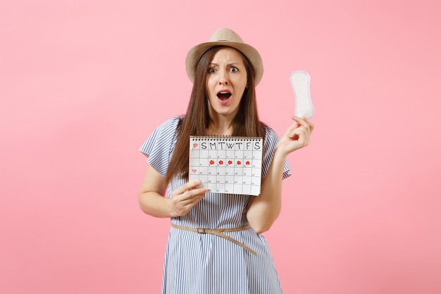 Femme triste choquée en robe bleue, chapeau tenant une serviette hygiénique, calendrier des périodes féminines pour vérifier les jours de menstruation isolés sur fond rose. Concept médical, sanitaire, gynécologique. Espace de copie