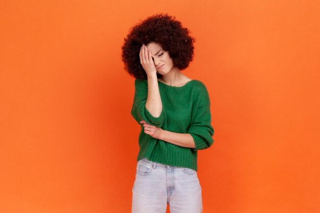 Femme triste avec une coiffure afro portant un chandail vert faisant un geste facepalm gardant la main sur la tête, se blâmant pour la mauvaise mémoire, erreur impardonnable. Studio intérieur tourné isolé sur fond orange