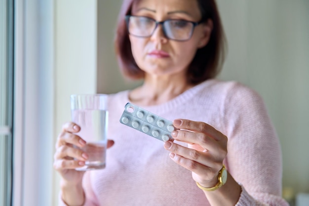 Femme triste mature avec blister de pilules et verre d'eau à la maison