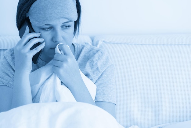 Femme triste portant un masque de sommeil à l'aide de smartphone alors qu'elle est assise dans son lit recouvert de couette