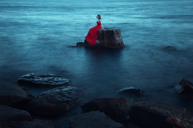 Une femme triste en robe rouge près de la mer en soirée. Exposition longue durée