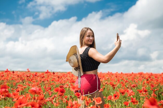 Femme ukrainienne en vêtements de sport et chapeau de paille prenant une photo selfie avec smartphone dans le champ de coquelicots en journée d'été.