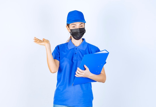 Femme en uniforme bleu et masque noir tenant un dossier bleu et pointant quelqu'un de côté.