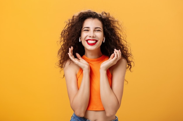 Femme urbaine séduisante et élégante avec une coiffure frisée et un anneau de nez souriant largement se sentant ravie d'écouter la piste préférée dans des écouteurs sans fil levant les paumes près du visage posant sur un mur orange.