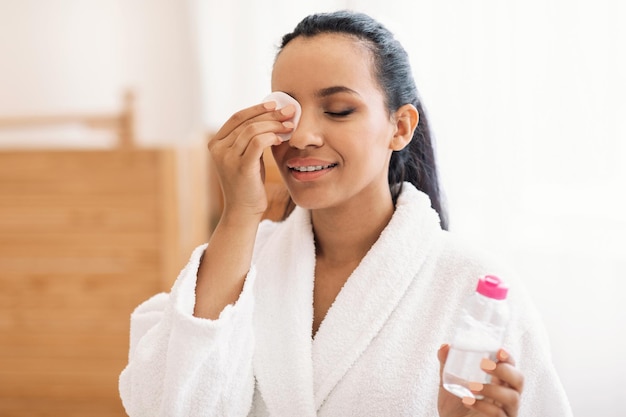 Femme utilisant de l'eau micellaire et un tampon de coton nettoyant le visage à l'intérieur