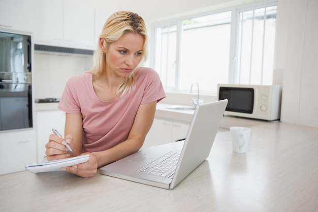 Femme utilisant un ordinateur portable tout en écrivant des notes dans la cuisine