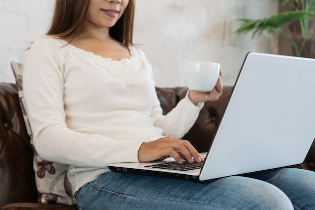 Femme utilisant un ordinateur portable tout en tenant une tasse de café et assis sur un canapé dans le salon.