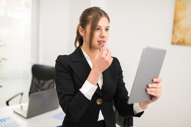 Femme utilisant sa tablette comme un miroir pour mettre du rouge à lèvres dans le bureau