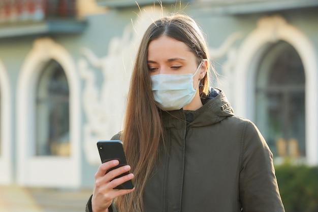 Une femme utilisant un smartphone porte un masque médical pour éviter la propagation du coronavirus dans une rue. Une fille avec un masque chirurgical sur le visage contre COVID-19.