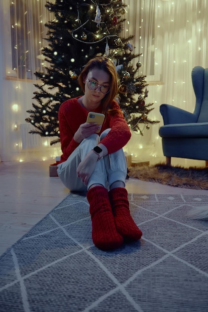 Femme utilisant un téléphone portable assis à côté de l'arbre de Noël sur le sol à la maison