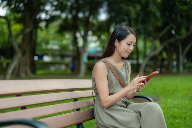 Une femme utilise un smartphone et s'assoit sur un banc dans le parc.