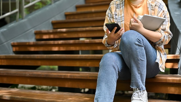 Une Femme Utilise Son Smartphone Alors Qu'elle Est Assise Dans Les Escaliers De L'image Recadrée Du Parc De Son Campus