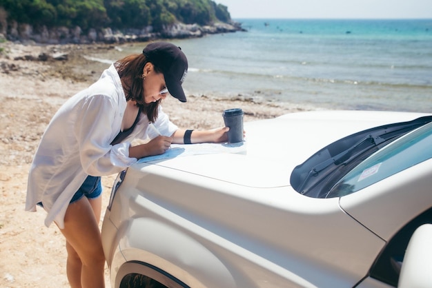 Femme vérifiant la carte sur le capot de la voiture en buvant du café sur la plage de la mer d'été