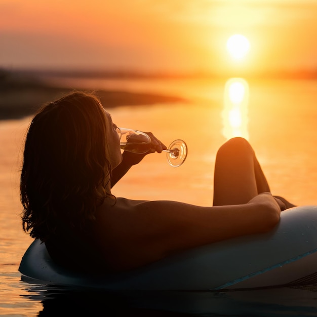 Femme avec un verre de champagne allongé sur un anneau gonflable dans l'eau au coucher ou au lever du soleil