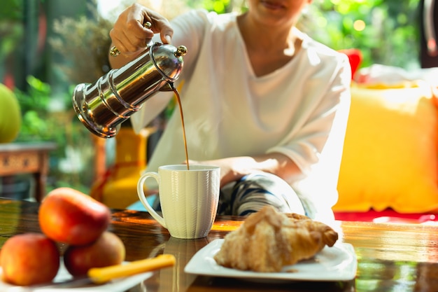 Femme versant du café dans une tasse d'une cafetière avec de la nourriture sur la table.