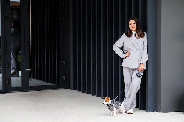 Femme en veste décontractée debout avec son chien Jack Russell Terrier de race pure moelleux préféré habillé en costume pour chiens près du mur en plein air le jour de l'automne