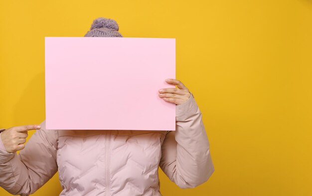 Femme en veste d'hiver rose et chapeau tient une feuille de papier rose vierge sur fond jaune. Vente saisonnière. Lieu d'inscription