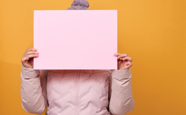 Femme en veste d'hiver rose et chapeau tient une feuille de papier rose vierge sur fond jaune. Vente saisonnière. Lieu d'inscription