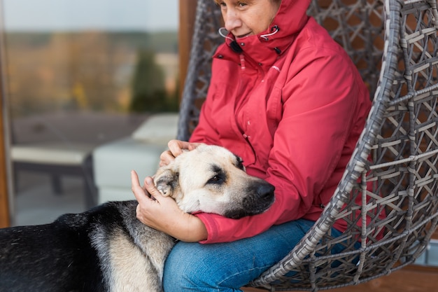 une femme en veste rouge joue et caresse un chien de berger assis sur une chaise en osier