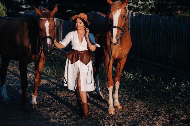 Une femme en vêtements blancs et un chapeau marche avec des chevaux dans la nature