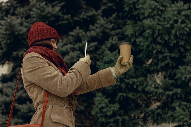 femme en vêtements chauds prenant une photo d'une tasse de papier vierge avec du café à emporter sur smartphone