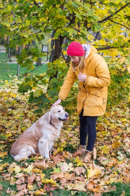 Femme en vêtements chauds avec son retriever dans un parc d'automne parmi les feuilles jaunes.