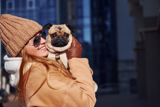 Une femme en vêtements chauds tient son petit chien carlin sur les mains près de l'immeuble d'affaires qui se trouve en arrière-plan.