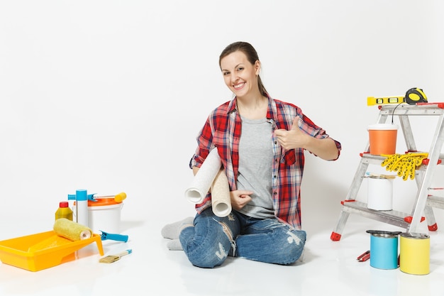 Femme en vêtements décontractés assis sur le sol avec des rouleaux de papier peint, instruments pour appartement de rénovation isolé sur fond blanc. Accessoires pour le collage d'outils de peinture. Réparer le concept de maison. Espace de copie