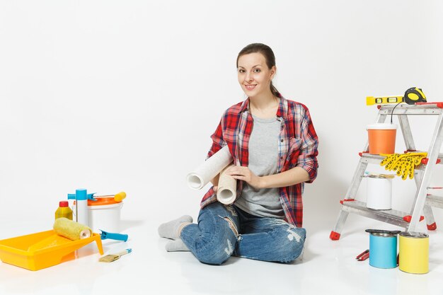 Femme en vêtements décontractés assis sur le sol avec des rouleaux de papier peint, instruments pour appartement de rénovation isolé sur fond blanc. Accessoires pour le collage d'outils de peinture. Réparer le concept de maison. Espace de copie