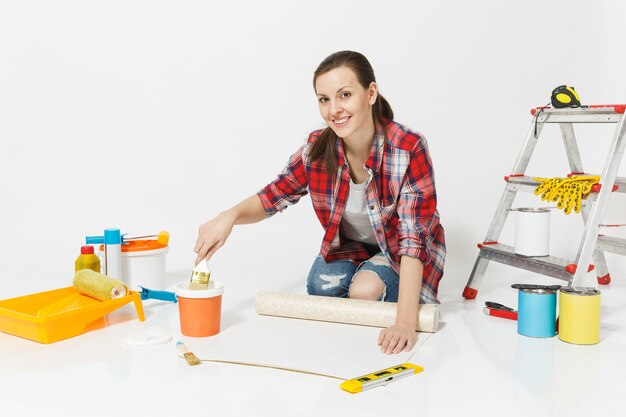 Femme en vêtements décontractés assise sur le sol avec des rouleaux de papier peint, une brosse, des instruments pour l'appartement de rénovation isolé sur fond blanc. Accessoires pour le collage, outils de peinture. Réparer le concept de maison.