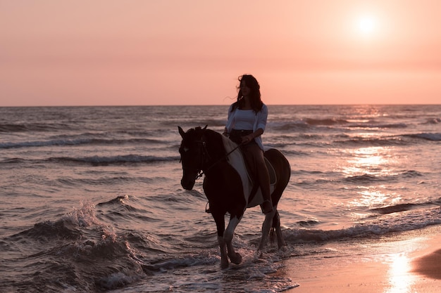 Femme en vêtements d'été aime monter à cheval sur une belle plage de sable au coucher du soleil mise au point sélective