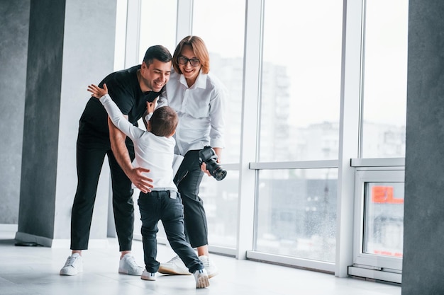 Femme en vêtements formels et avec caméra à la main debout à l'intérieur d'une pièce vide avec un homme et un petit garçon qui courent et s'amusent