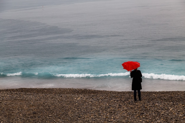 femme en vêtements noirs avec parapluie rouge debout seule sur la plage de galets et regardant la mer