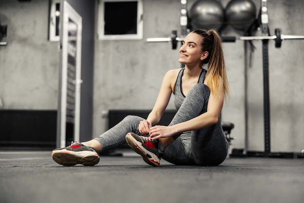 Une Femme En Vêtements De Sport Gris Est Assise Sur Le Sol D'une Salle De Sport Intérieure Et Se Prépare à Commencer L'entraînement