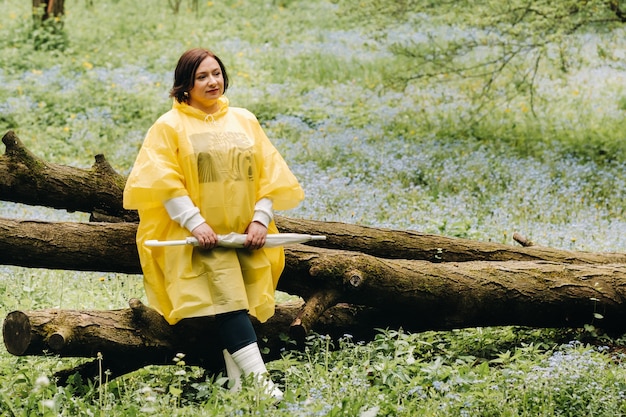 Une femme vêtue d'un imperméable jaune avec un parapluie est assise sur un arbre tombé dans la forêt en été.