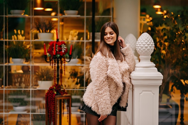 Une femme vêtue d'un manteau de fourrure rose se tient devant une devanture de magasin avec un étalage de fleurs.